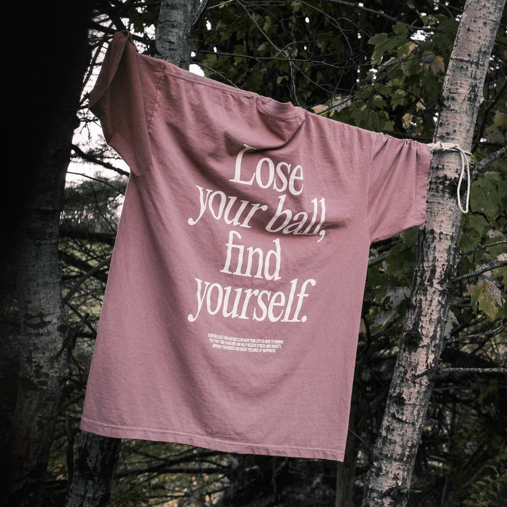 Pink t-shirt with text 'Lose your ball, find yourself' hanging on a tree branch in a forest setting.